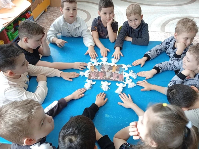 На фотографии изображена группа детей, сидящих в кругу на синем коврике или покрытии. Дети внимательно смотрят на центр круга..